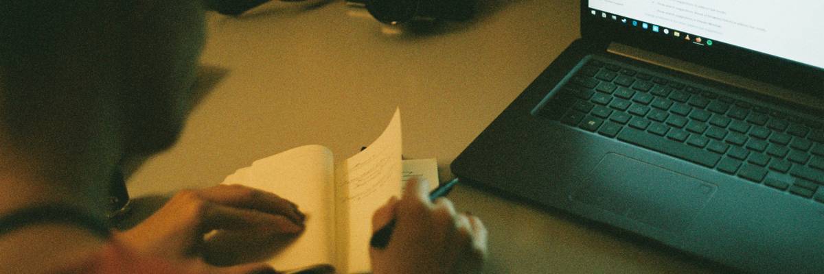 A person writing notes in a modern workspace, surrounded by digital productivity tools and a laptop displaying text analysis results